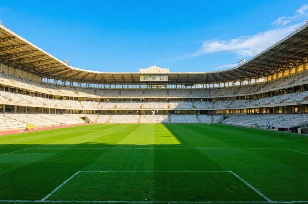 A lush green professional soccer field inside a historic open-roof stadium with empty grandstands under a bright blue sky